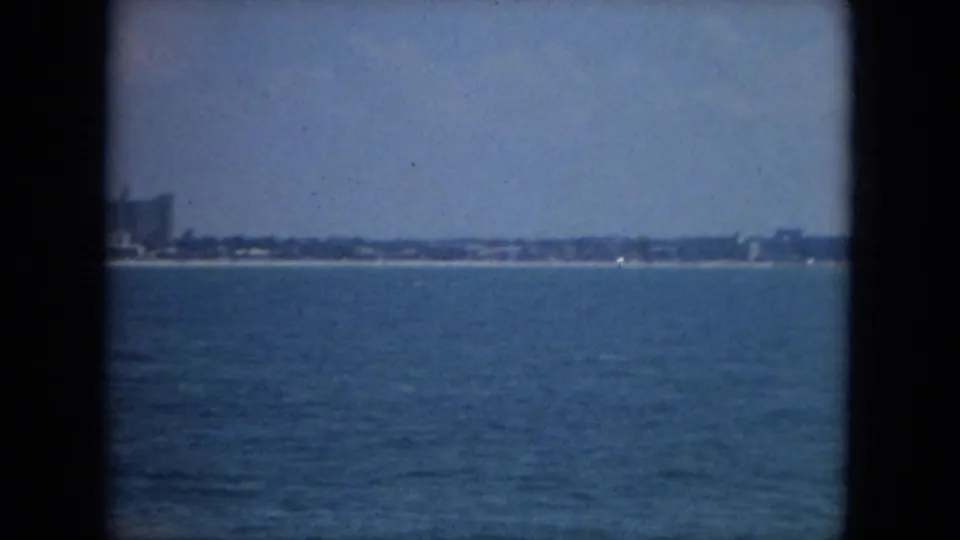 1967 Florida offshore view with blue water, a low coastal strip, distant buildings, and small boats near the horizon.