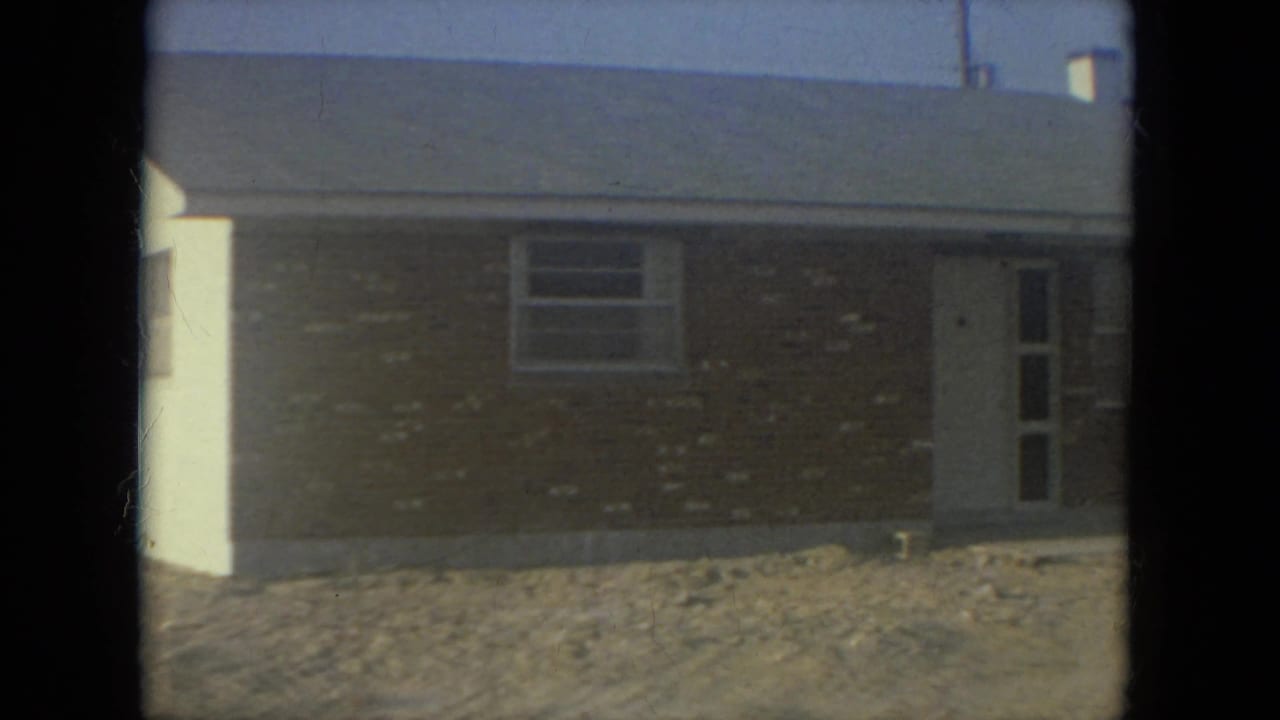 1967 Florida 8mm frame of a single-story brick house exterior with a white door, window, and sandy front yard.