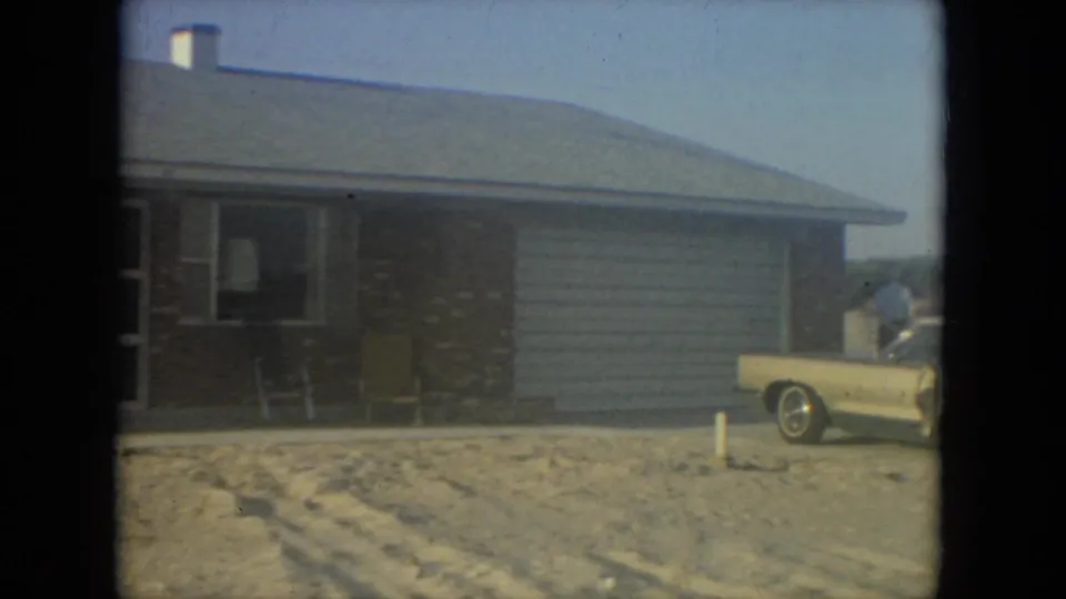 1967 Florida 8mm frame of a house exterior with porch chairs, sandy driveway, yellow car, and a person near the car.