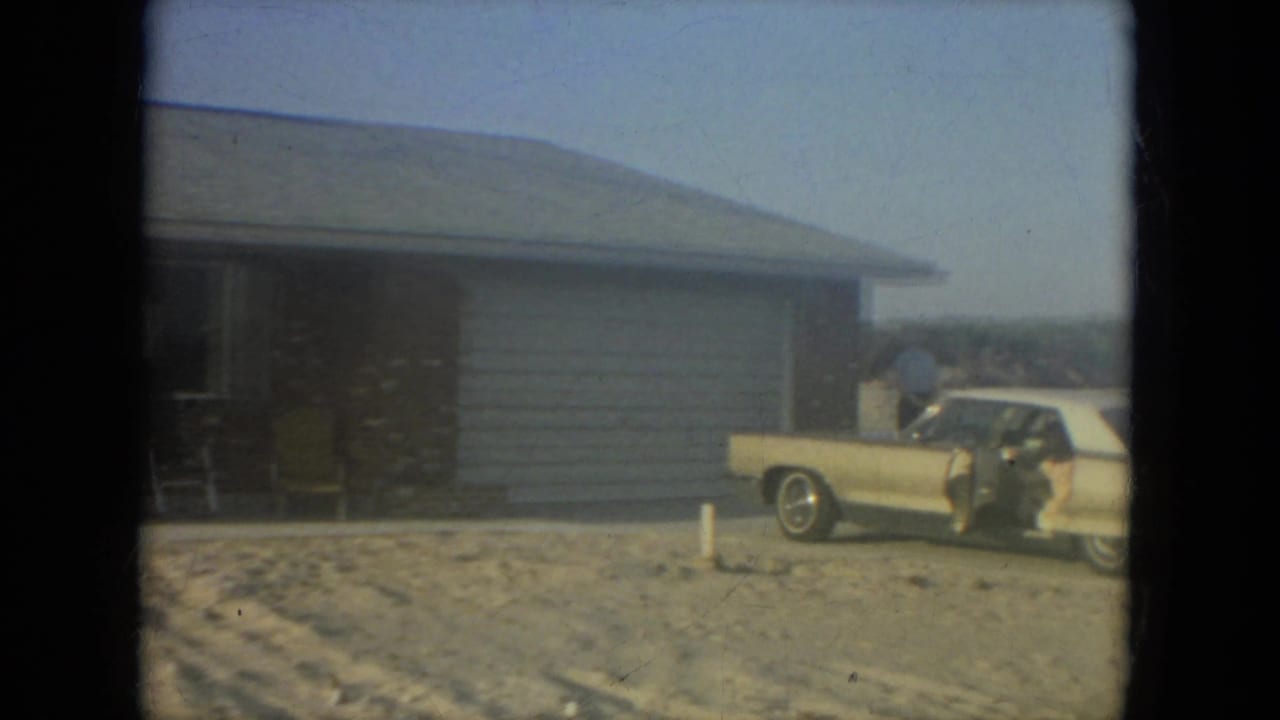1967 Florida home-movie frame showing a gold car by a brick ranch-style house with white siding and sandy yard.
