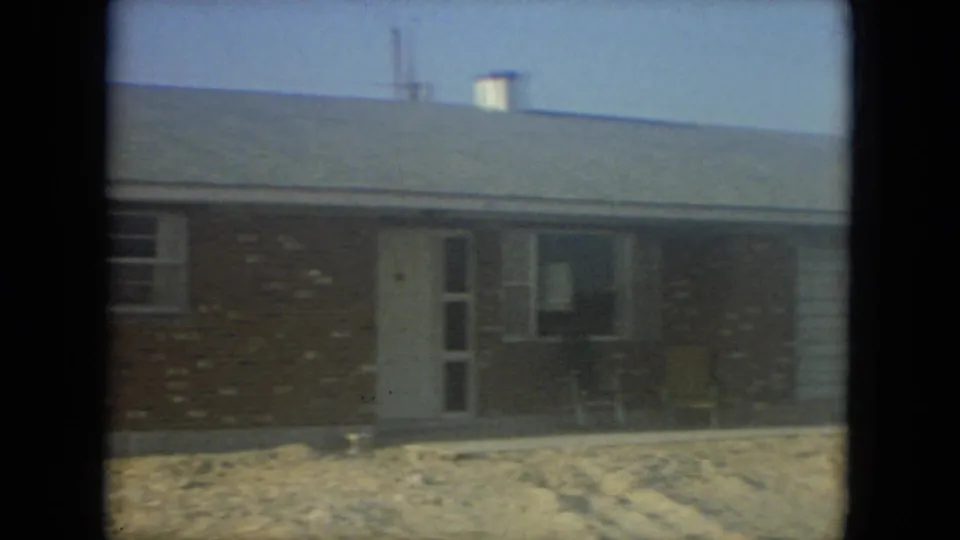 1967 Florida home-movie frame scanning the front entrance of a brick ranch house with porch chairs and sandy ground.