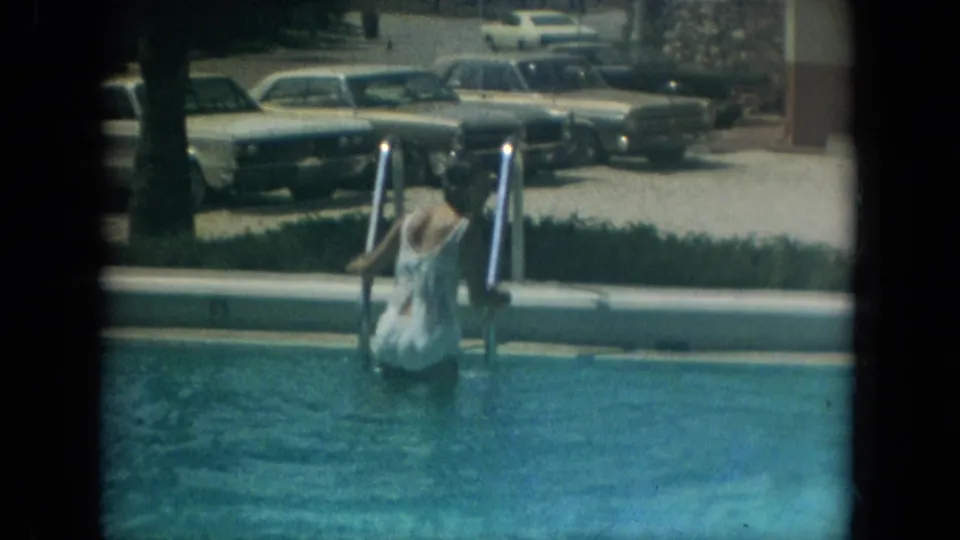 A woman in a white cover-up stands in the swimming pool holding ladder rails, with parked 1960s cars visible beyond the pool edge.