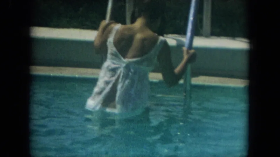 Closer 8mm view of the woman from behind as she holds the ladder rails and wades deeper into the resort pool.