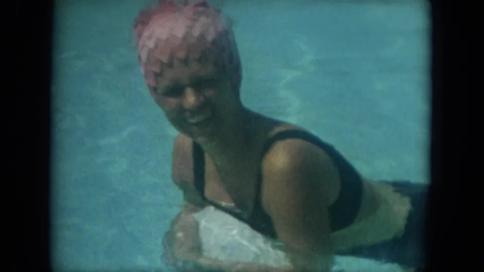 The woman, now wearing a pink swim cap and dark swimsuit under the white cover-up, smiles toward the camera in the pool.
