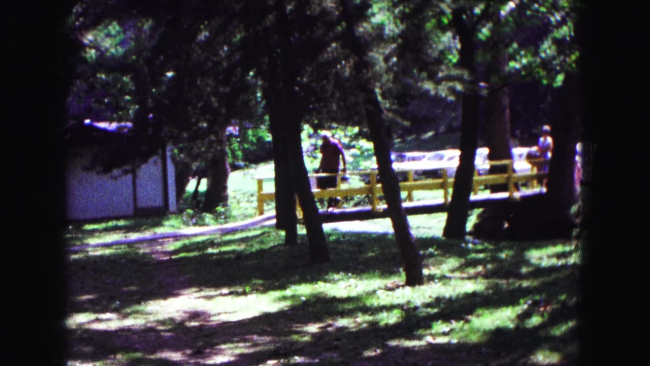 Shaded park path with trees, a white building, yellow railing, and distant people.