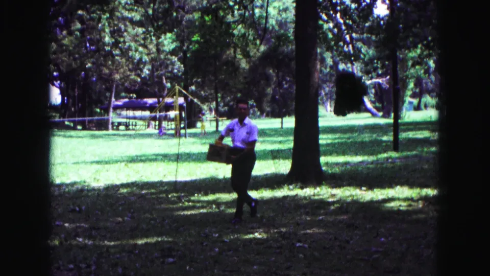 1967 Galena, Illinois home-movie frame of a man carrying a box across shaded grass, with trees and playground equipment in the background.