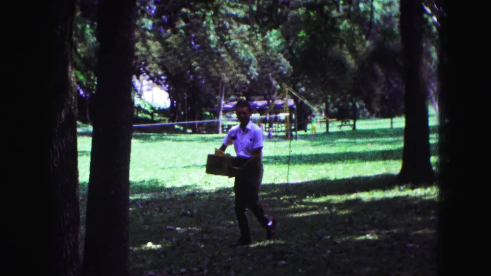1967 Galena, Illinois frame showing a man walking with a box through a shaded park, with trees, open grass, and distant play structures.
