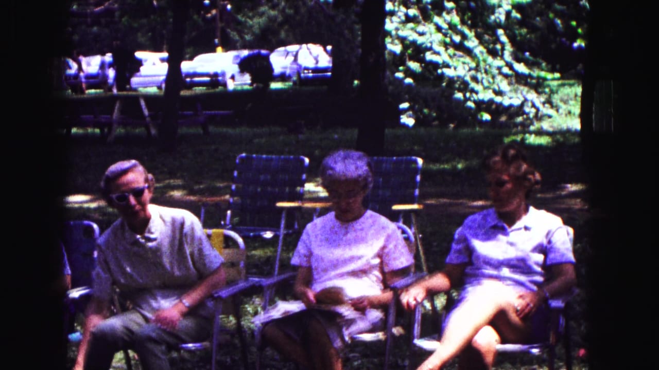 Three older adults sit in folding lawn chairs in a Galena park setting, with empty chairs and 1960s cars in the background.
