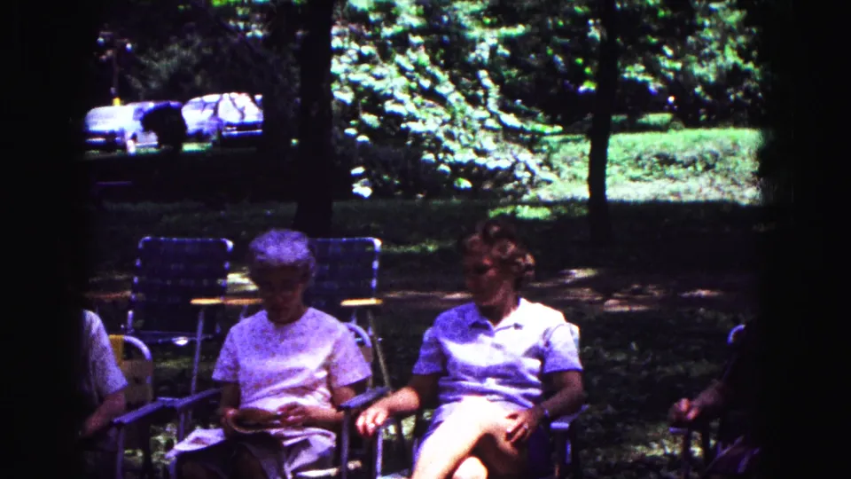 Two women sit in folding lawn chairs in the shaded 1967 Galena park scene, with trees and parked cars behind them.