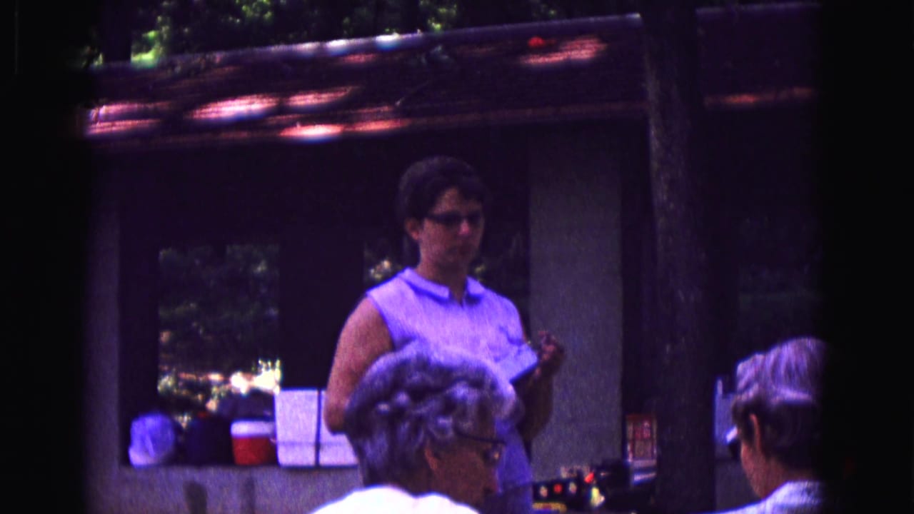 Woman in glasses stands by a counter while seated patrons face her in a dim restaurant interior.