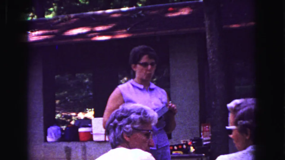 1967 8mm frame showing a woman in a sleeveless top behind seated restaurant patrons in likely Galena, Illinois.