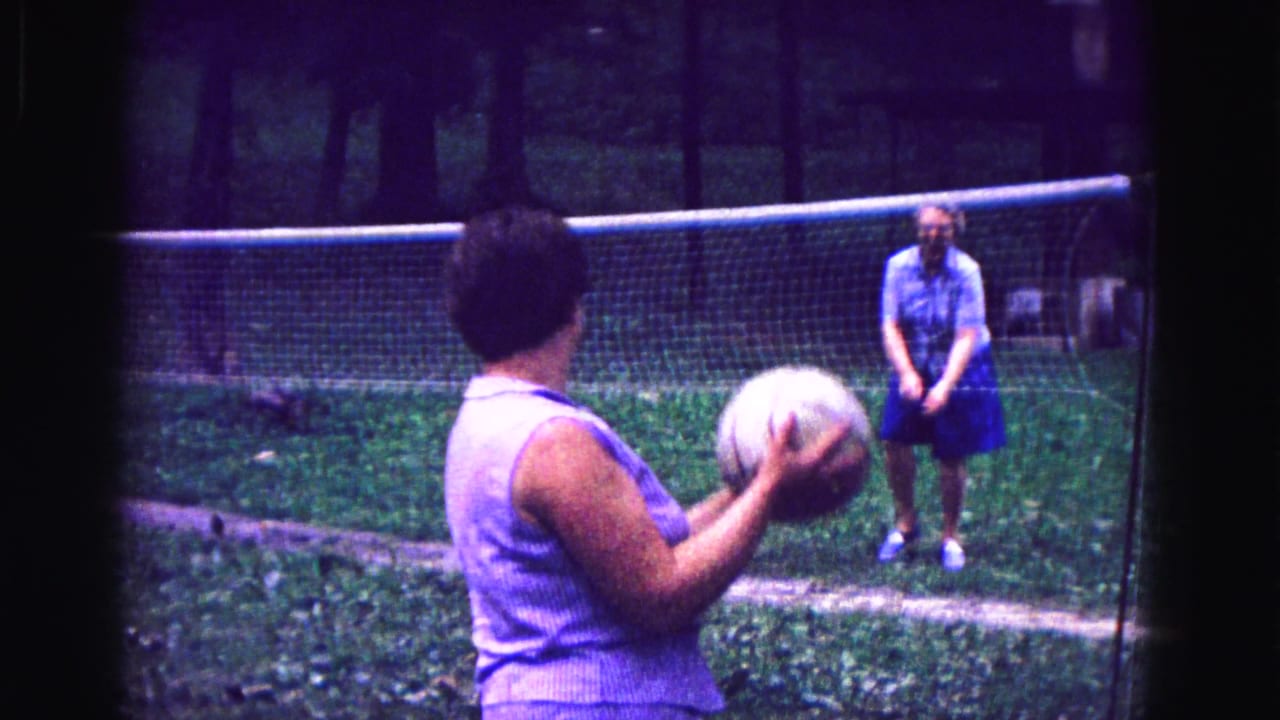 Woman holding a volleyball faces another woman across a backyard net.
