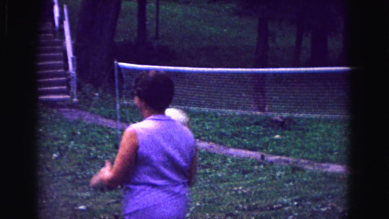 Woman seen from behind tosses a volleyball toward a backyard net.