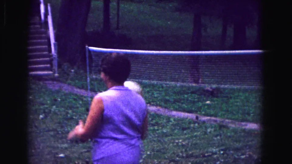 1967 Galena backyard volleyball frame showing a woman from behind as the ball moves toward the net in a shaded garden.
