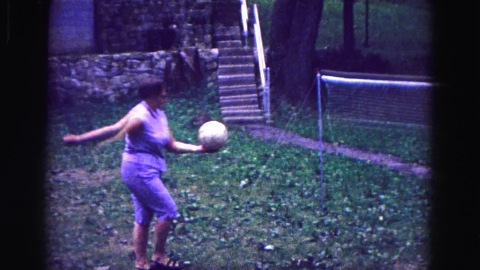 1967 Galena home-movie frame of a woman balancing or preparing the volleyball near a garden net with stone steps behind her.