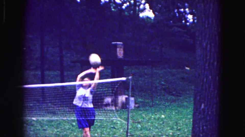 1967 Galena frame of a woman reaching up to meet the volleyball above a low net in a shaded grassy backyard.