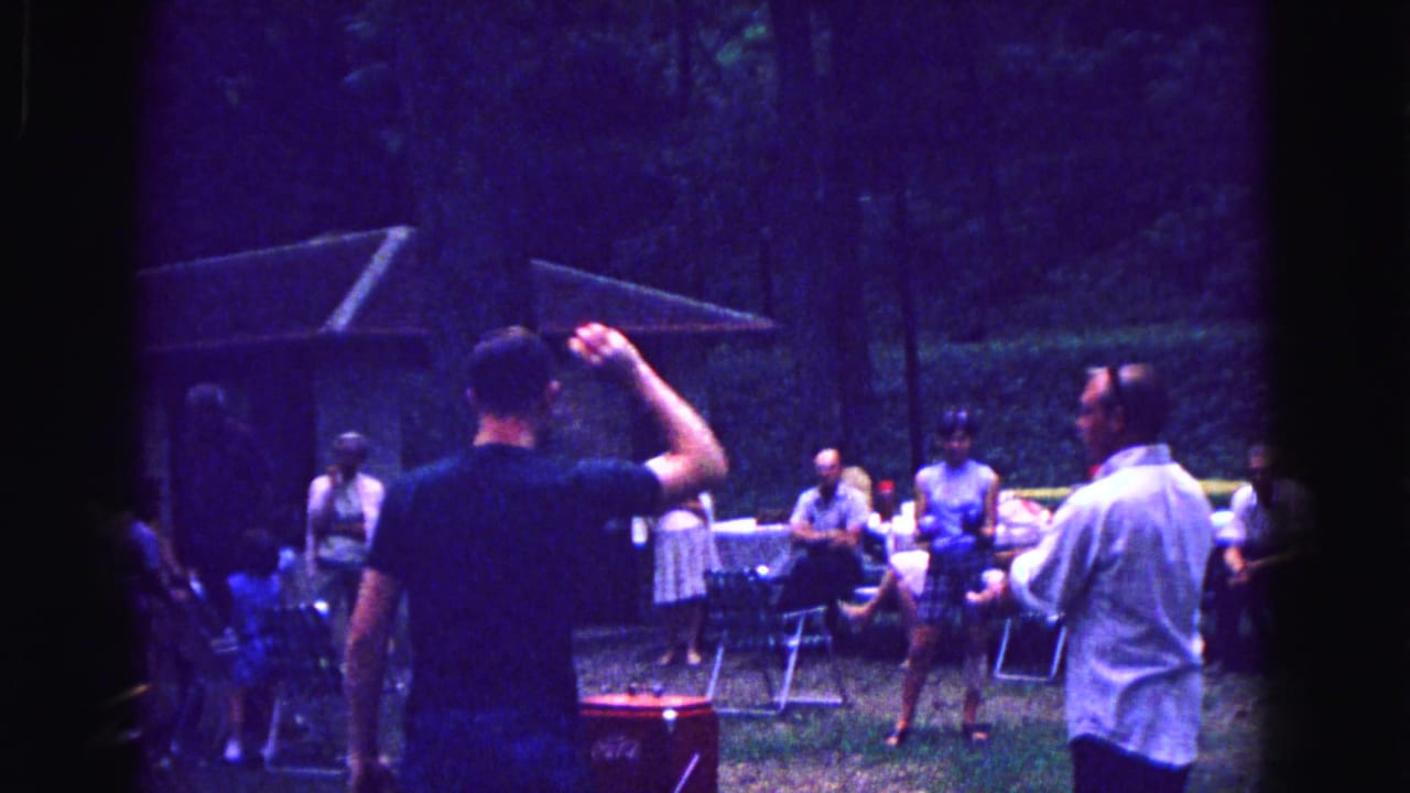 Adults gather near picnic tables and a red cooler in a wooded yard.