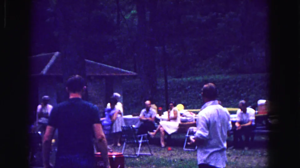 1967 Galena, Illinois home-movie frame showing a casual group gathered near folding chairs, picnic tables, and a red cooler under trees.