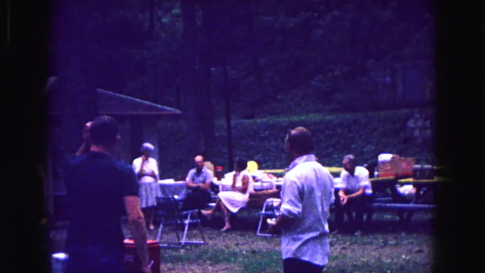 1967 Galena, Illinois home-movie frame of adults socializing around picnic tables and folding chairs in a shaded outdoor gathering area.