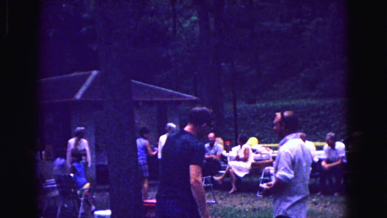 1967 home-movie frame from Galena, Illinois showing adults socializing at picnic tables beneath trees in a backyard.