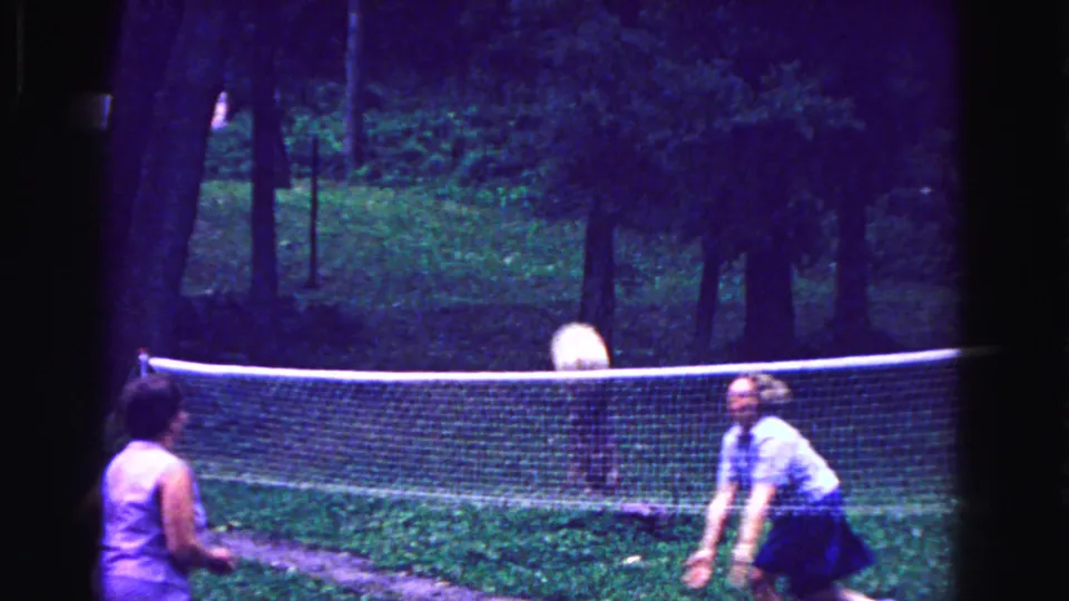 1967 Galena, Illinois frame with two adults playing volleyball across a backyard net under trees.