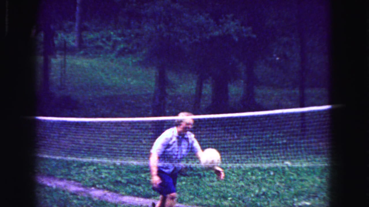 Woman on the far side of a net moves toward a volleyball in a yard.