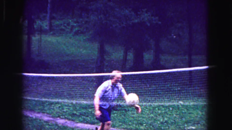 A woman in a patterned top and dark skirt approaches the volleyball beside a backyard net in the 1967 Galena home movie.
