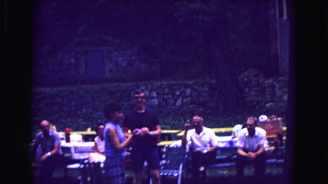 1967 Galena, Illinois home-movie frame showing adults gathered at picnic tables beneath trees, with a low stone wall in the background.