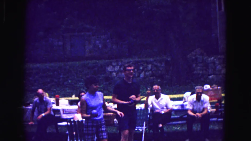 1967 Galena home-movie frame showing adults beside picnic tables, with a woman in a blue top near a man in dark clothing.