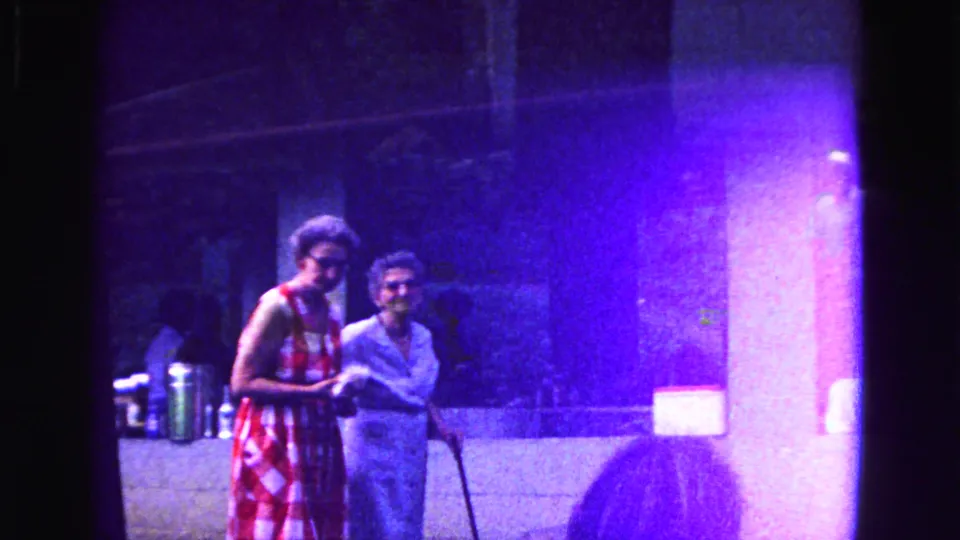 1967 Galena home-movie frame showing two women near a building, one in a red and white checkered dress and another with a cane.