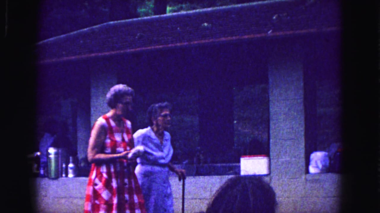 Two elderly women walk arm in arm near a stone shelter, one holding a cane.