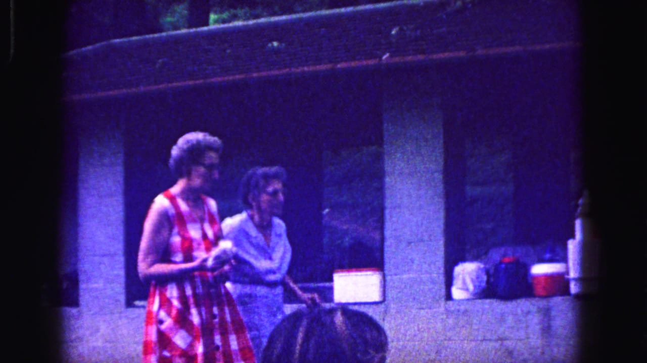Two elderly women in 1967 Galena stand arm in arm by a stone shelter with picnic coolers and bags nearby.