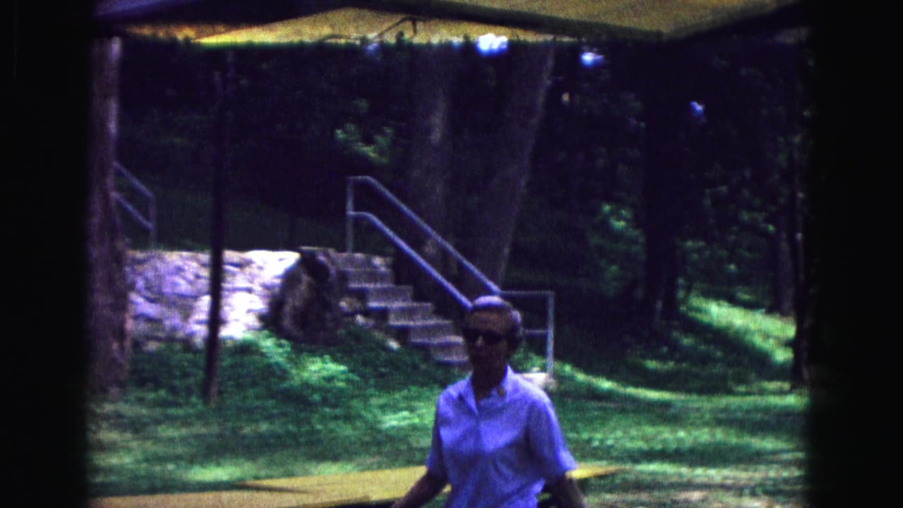Woman in a light shirt walks near steps, a metal railing, trees, and stone walls.
