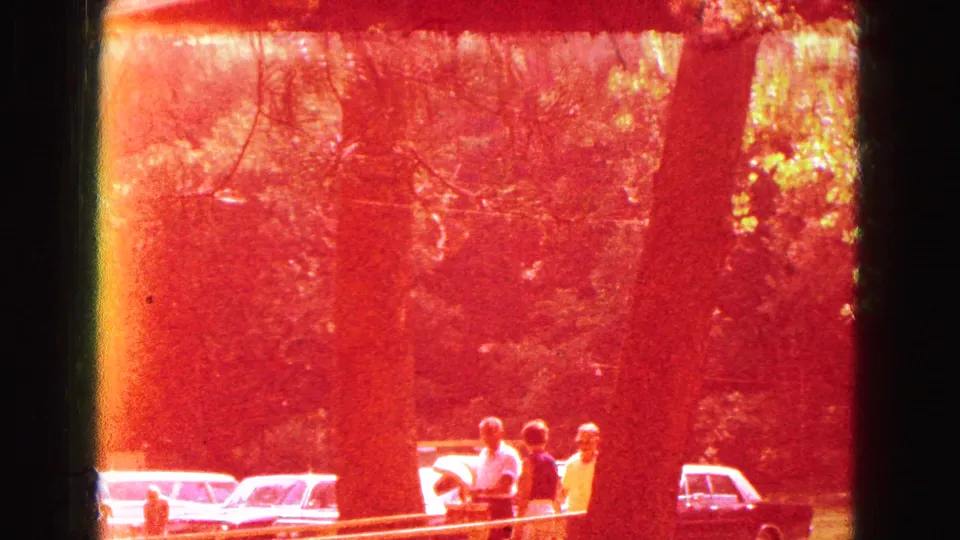 A red color shift covers a park scene with several people near picnic tables, large trees, and parked cars in the background.