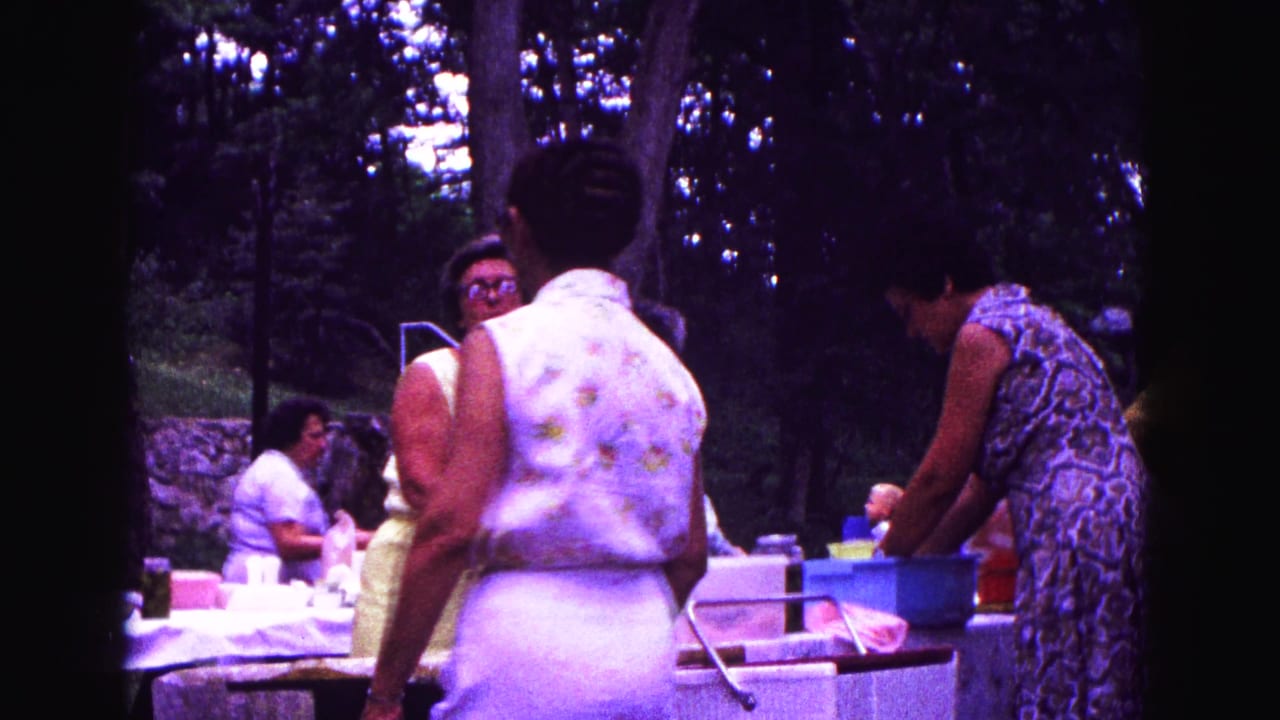 Women gather around an outdoor picnic table with containers in a wooded setting.