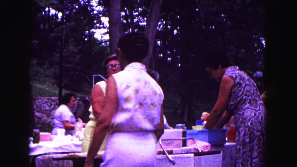 1967 Galena, Illinois home-movie frame showing women near a picnic table with food containers and trees behind them.