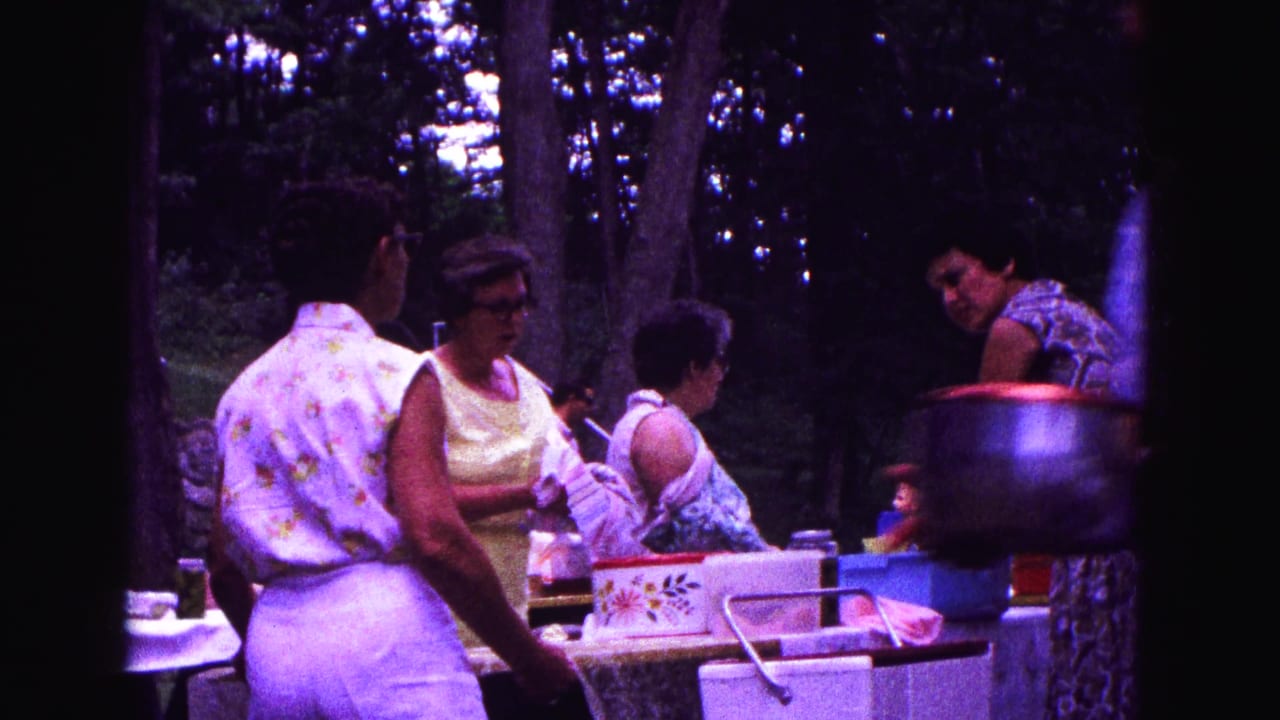 1967 Galena, Illinois frame with women preparing picnic items beside a table, blue basin, patterned box, and large pot.