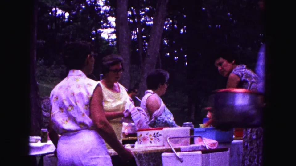 1967 Galena, Illinois frame with women preparing picnic items beside a table, blue basin, patterned box, and large pot.