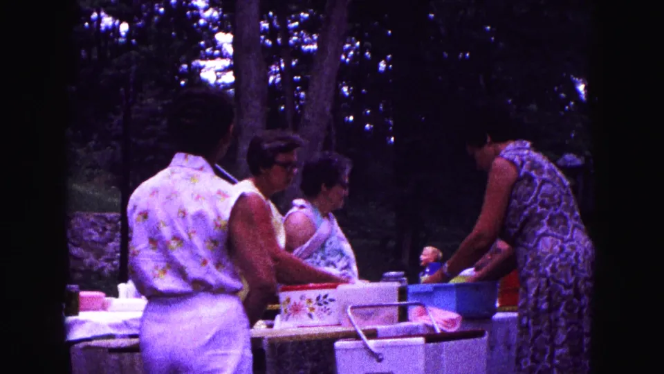1967 Galena, Illinois home-movie frame showing women arranging picnic containers at a table under trees.