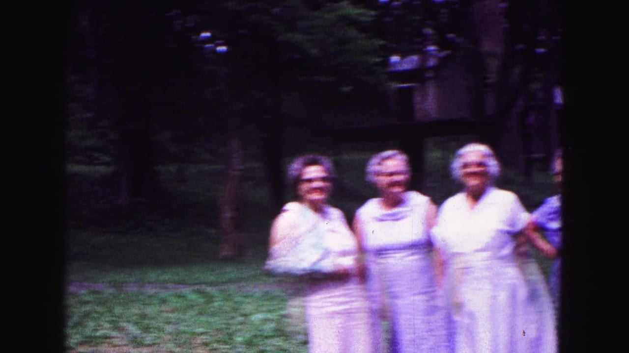 Three older women in light dresses pose on a shaded lawn.