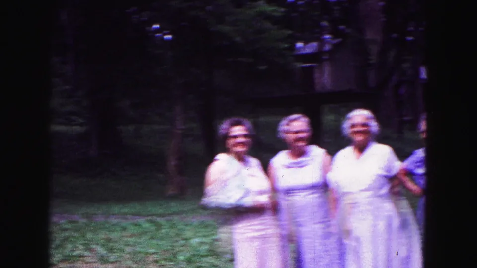 Three older women pose together on a shaded Galena lawn in 1967 home-movie footage, with trees and grass behind them.