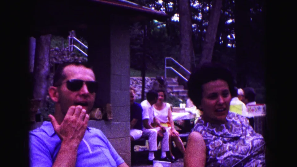 A man in sunglasses and a woman stand near picnic tables during the 1967 Galena backyard gathering, with seated guests in the background.