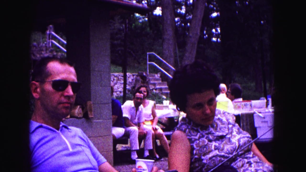 Seated man in sunglasses and woman in patterned dress with people and picnic tables behind them.