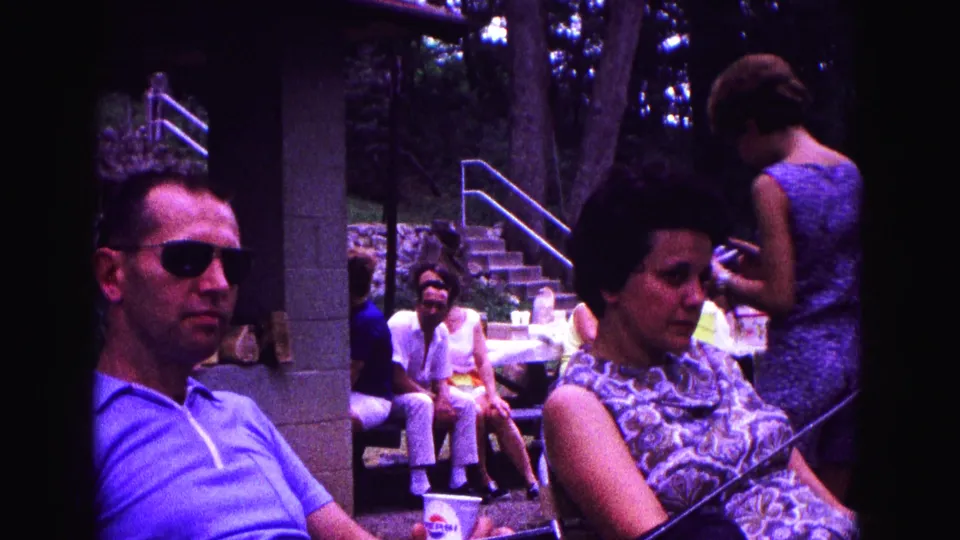 At the park gathering, a man in sunglasses and a woman in a patterned dress sit while another adult stands nearby beside picnic tables.