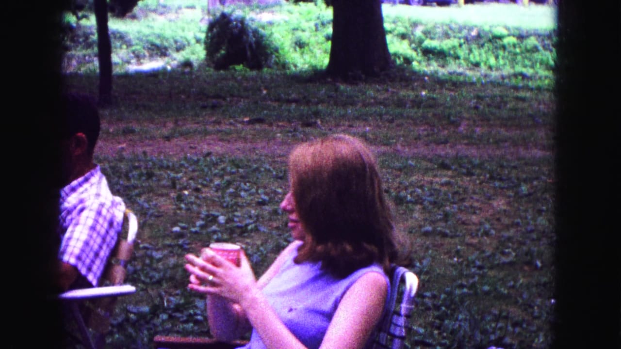 A young woman in a purple dress holds a red cup during a 1967 Galena outdoor gathering, with a plaid-shirted man nearby.