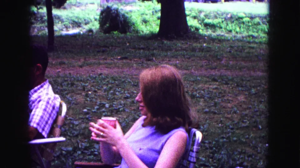 A young woman in a purple dress holds a red cup during a 1967 Galena outdoor gathering, with a plaid-shirted man nearby.