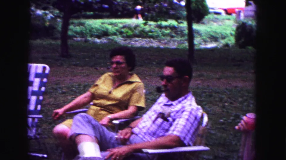 Adults relax in folding chairs in a grassy Galena yard, with trees and parked cars visible in the 1967 home-movie frame.