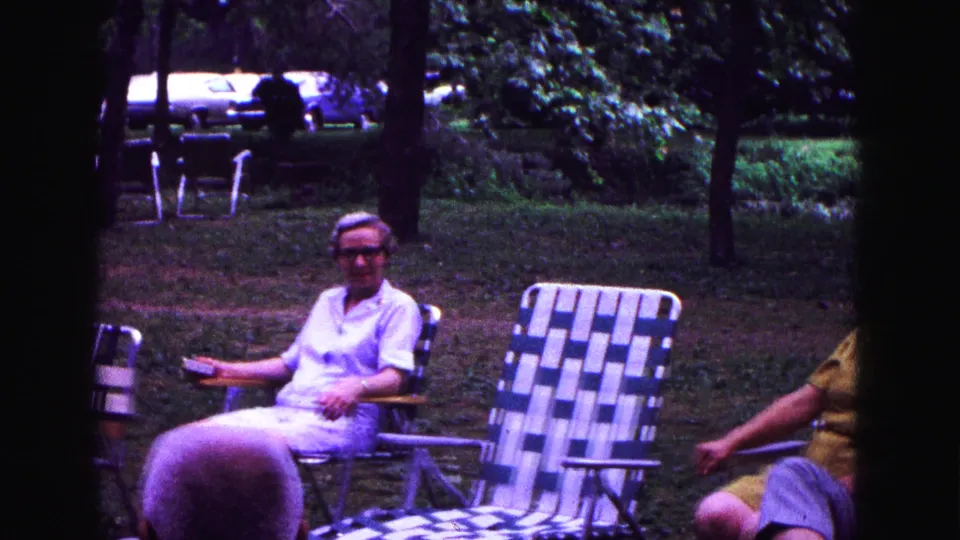 An older woman sits in a lawn chair in the Galena yard, with an empty striped chair, trees, grass, and parked cars behind her.