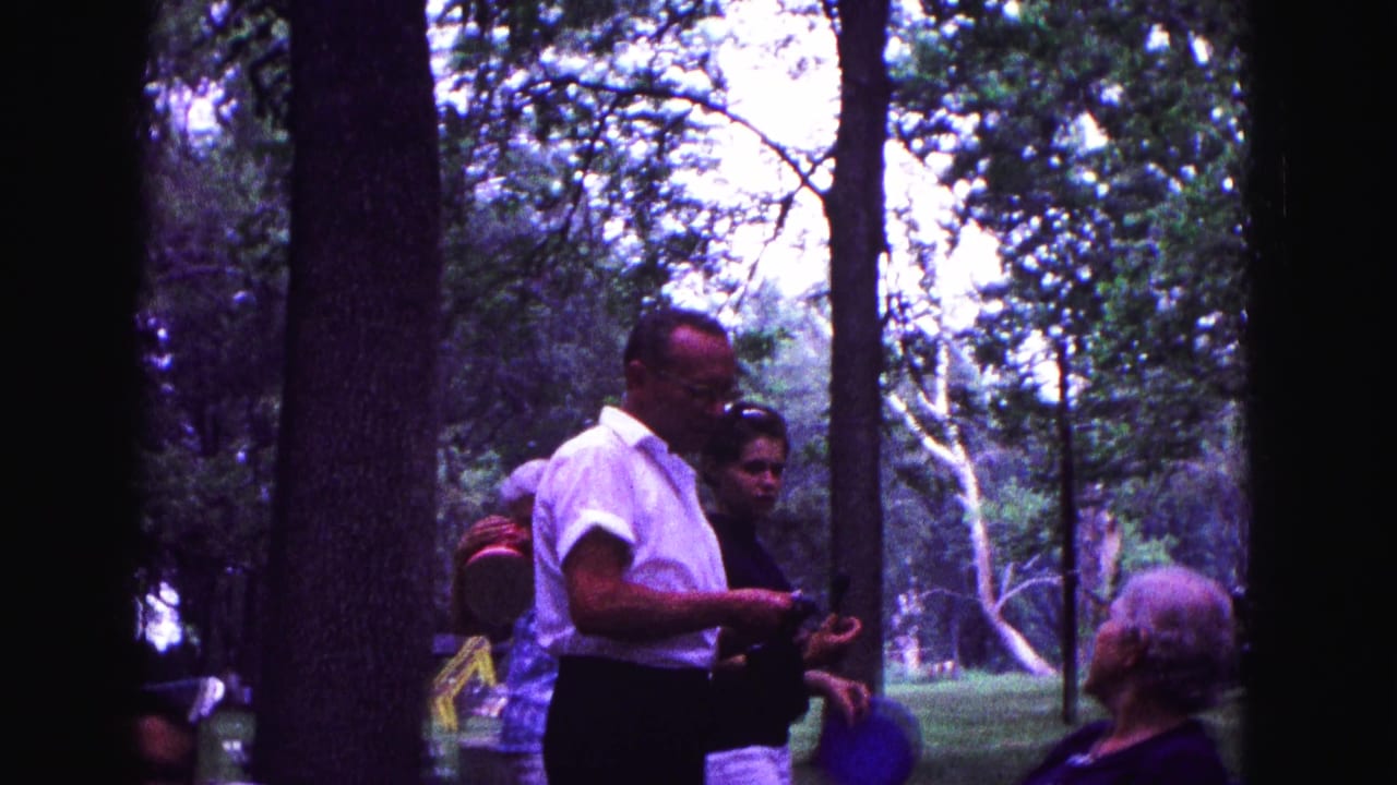 Adults gather under trees while a man in a white shirt holds a camera.