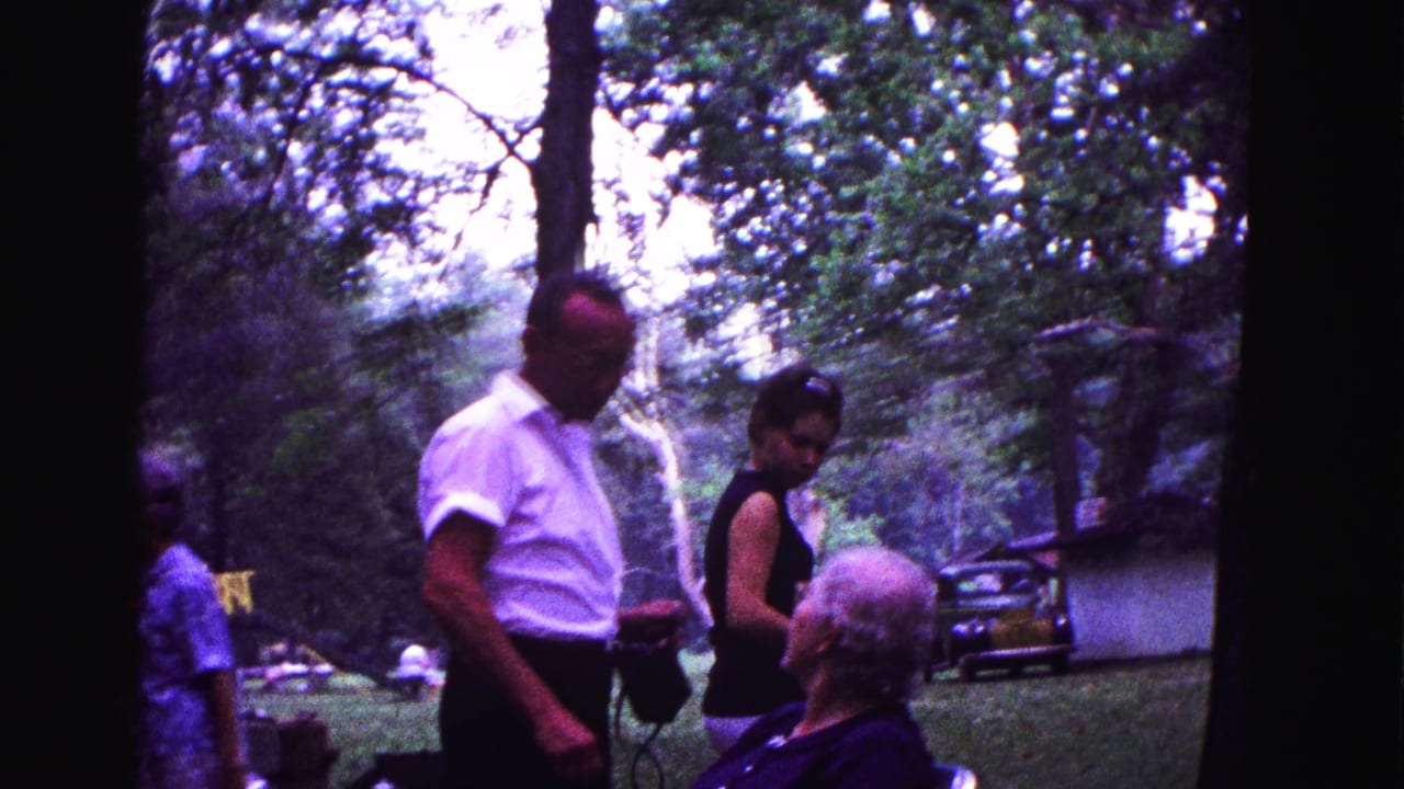 A man with a camera stands near seated and standing adults in a wooded park.